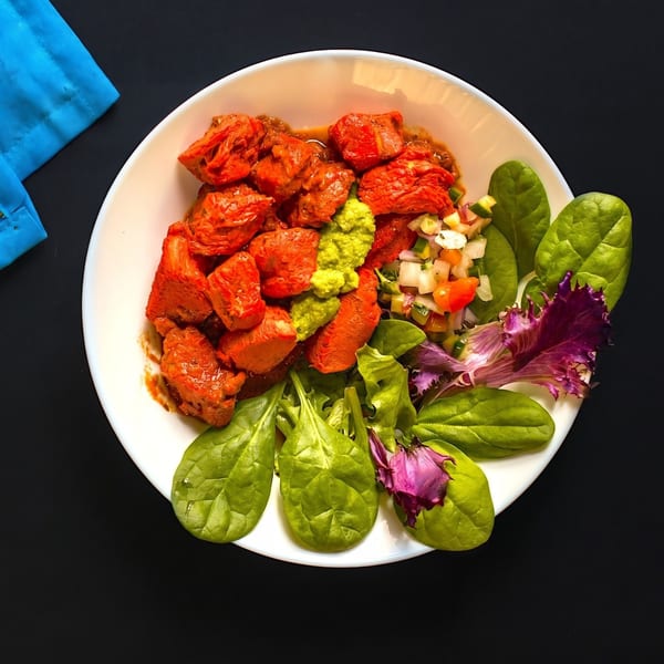 Top-down view of a white bowl filled with red chicken pieces, guacamole, salsa, and spinach.